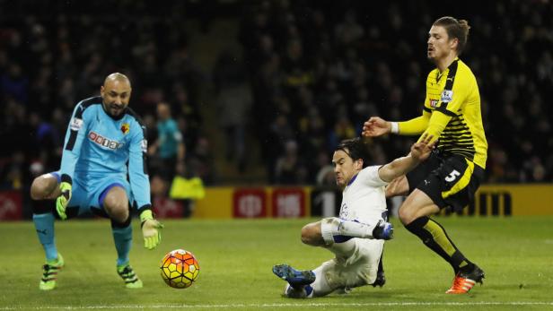 Ein Fußballspieler grätscht nach dem Ball, während ein Torwart und ein anderer Spieler zusehen.