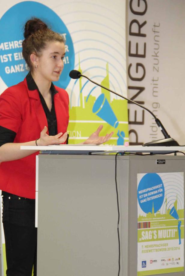 Eine junge Frau hält eine Rede auf einem Podium mit dem Logo „Sag's Multi!“.