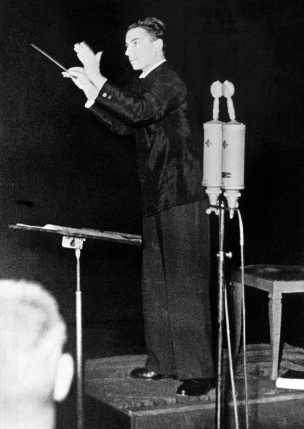 Ein Dirigent steht vor einem Notenständer und dirigiert ein Orchester.