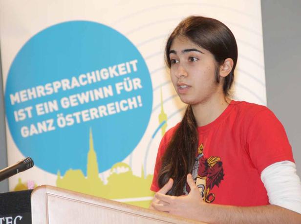 Eine junge Frau spricht an einem Rednerpult vor einem Hintergrund mit der Aufschrift „Mehrsprachigkeit ist ein Gewinn für ganz Österreich!“.