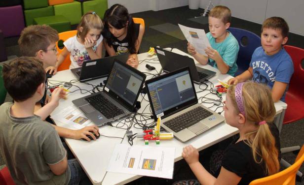 Eine Gruppe Kinder sitzt an einem Tisch und arbeitet mit Laptops und Robotik-Bausätzen.