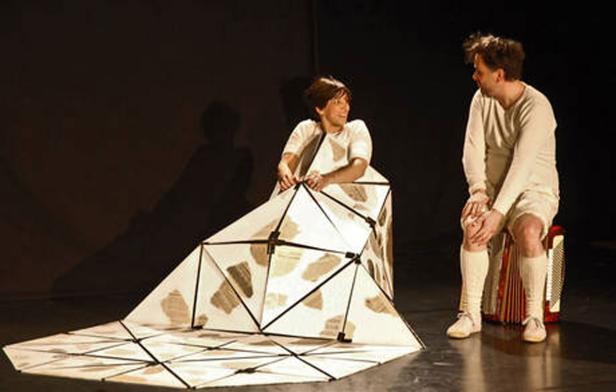 Zwei Schauspieler auf einer Bühne, eine Frau in einem geometrischen Kostüm, ein Mann sitzt auf einem Akkordeon.
