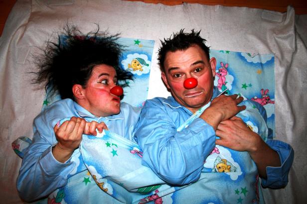 Zwei Clowns mit roter Nase liegen in blauen Schlafanzügen im Bett.