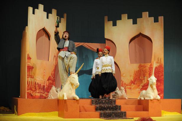 Eine Bühnenszene mit einem Mann, der eine Flasche hochhält, und zwei Frauen in Uniformen vor einer gemalten Burgkulisse.