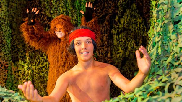 Ein junger Mann mit rotem Stirnband und ein Bär stehen vor einem grünen Hintergrund.