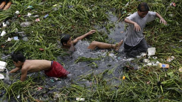 Drei Jungen spielen in einem verschmutzten Fluss zwischen Müll und Vegetation.