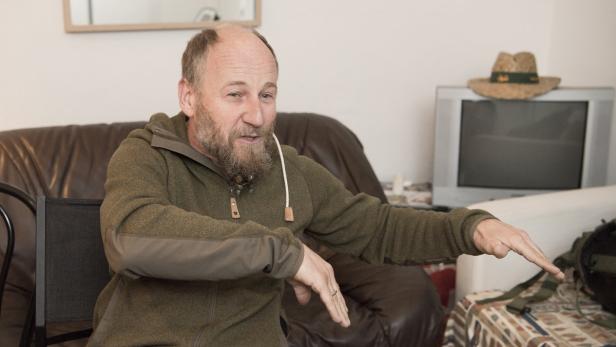 Ein Mann mit Bart gestikuliert, während er auf einem braunen Ledersofa sitzt.