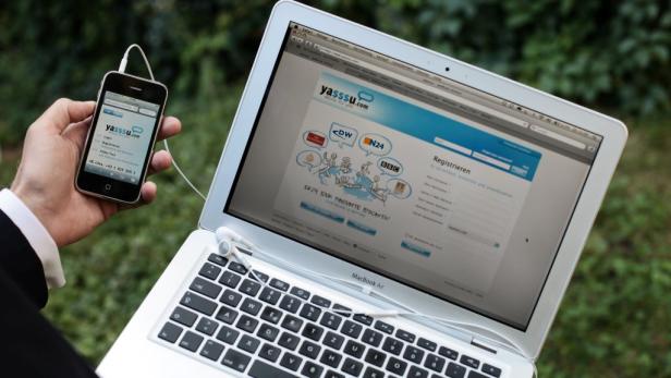 Ein Mann hält ein iPhone mit der Webseite yasssu.com neben einem MacBook Air.