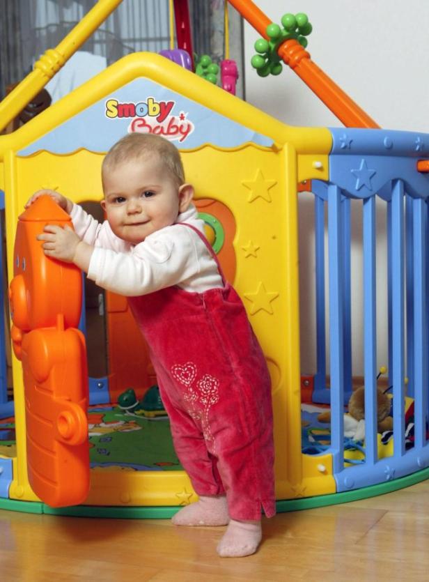 Ein Baby in rotem Overall steht an einem bunten Spielhaus von Smoby.