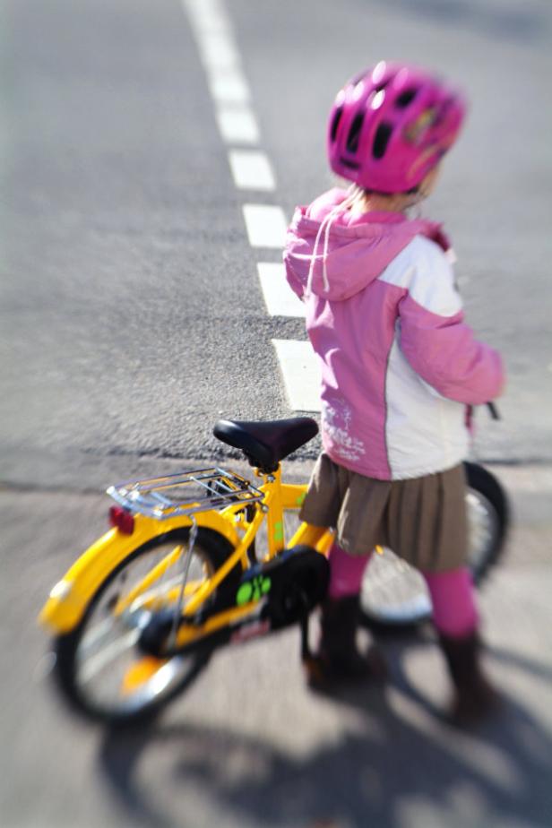 Ein kleines Mädchen mit rosa Helm steht neben einem gelben Fahrrad auf der Straße.