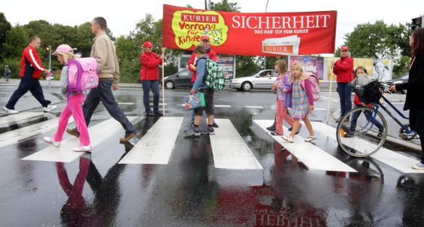 Kinder überqueren mit Unterstützung von Erwachsenen einen Zebrastreifen vor einem „Kurier“-Sicherheitsbanner.