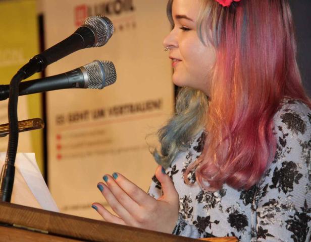 Eine junge Frau mit bunten Haaren spricht an einem Rednerpult vor zwei Mikrofonen.