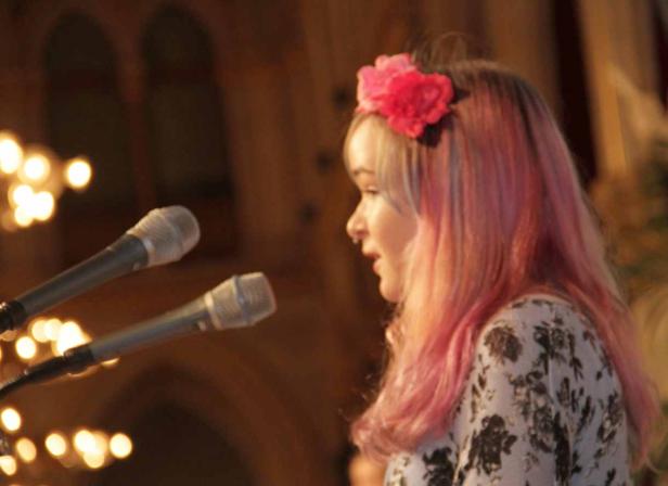 Eine junge Frau mit rosa Haaren und Blumenkranz steht vor zwei Mikrofonen.