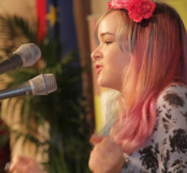 Eine junge Frau mit Blumen im Haar spricht vor zwei Mikrofonen.