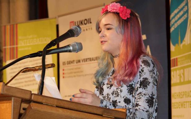 Eine junge Frau mit Blumenkranz hält eine Rede an einem Rednerpult.