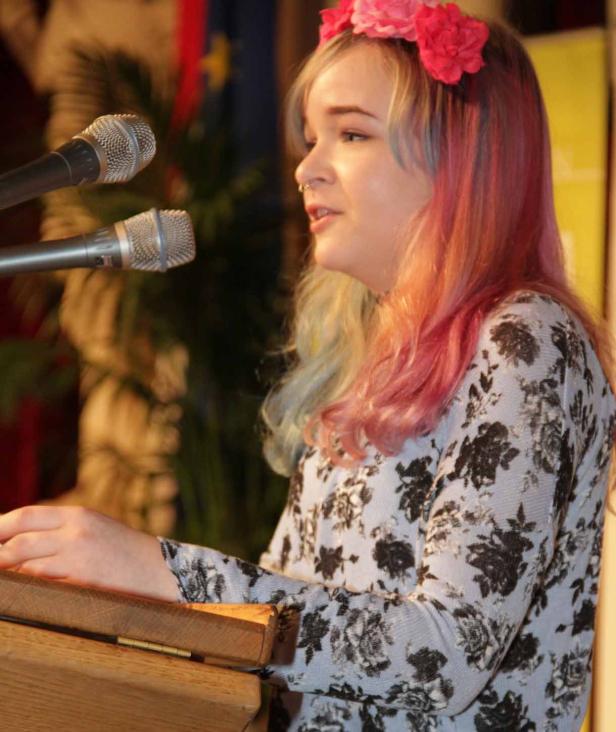 Eine junge Frau mit Blumenkranz im Haar spricht an einem Rednerpult.