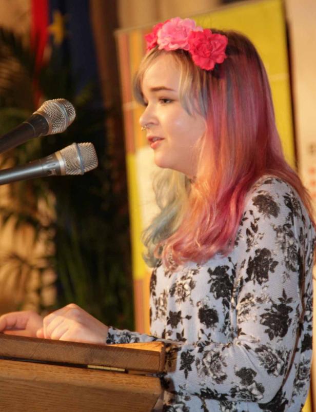 Eine junge Frau mit Blumenkranz hält eine Rede an einem Rednerpult.