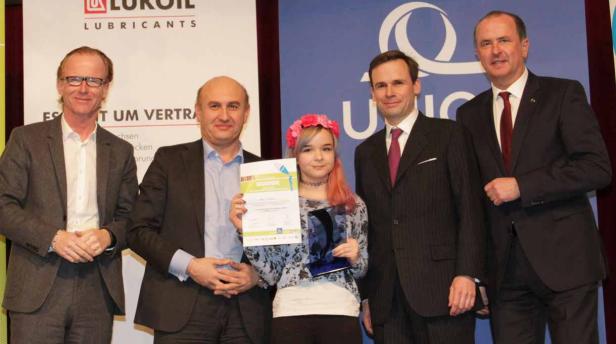 Eine junge Frau mit Blumenkranz hält eine Urkunde und einen Preis, umgeben von vier Männern in Anzügen.