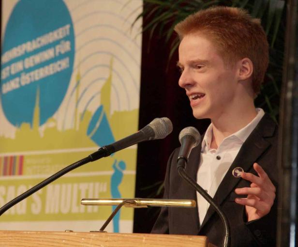 Ein junger Mann hält eine Rede vor Mikrofonen auf einer Bühne.