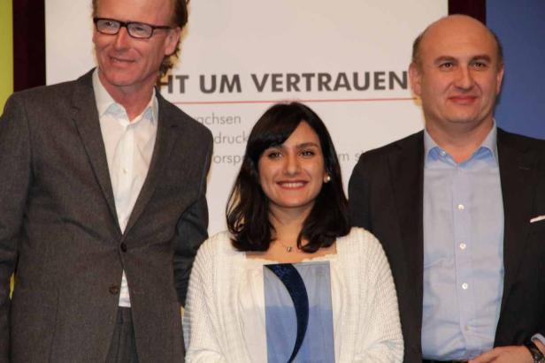 Drei Personen posieren vor einem Hintergrund mit dem Schriftzug „HT UM VERTRAUEN“.