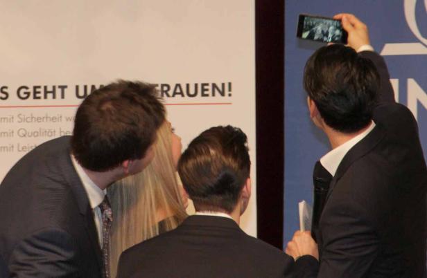 Drei Personen betrachten eine Präsentation, eine filmt sie mit dem Smartphone.