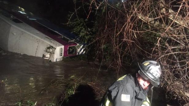 Ein entgleister Zug liegt teilweise im Wasser, während ein Feuerwehrmann im Vordergrund steht.