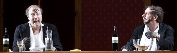 Zwei Männer mit schwarzer Farbe im Gesicht sitzen an einem Tisch mit Weinflaschen.