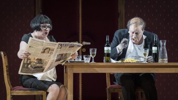 Eine Frau liest Zeitung, während ein Mann am Tisch isst.