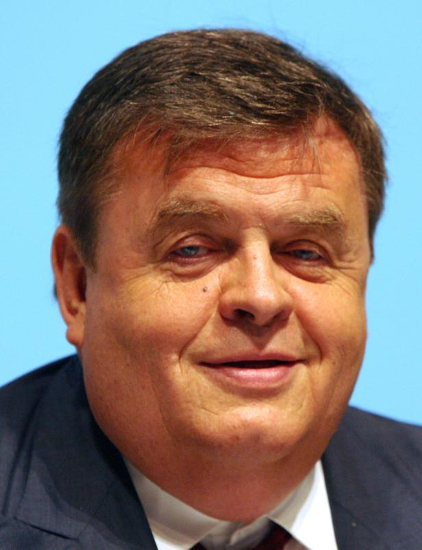 Ein Porträt eines Mannes mit braunen Haaren und einem dunklen Anzug vor einem hellblauen Hintergrund.