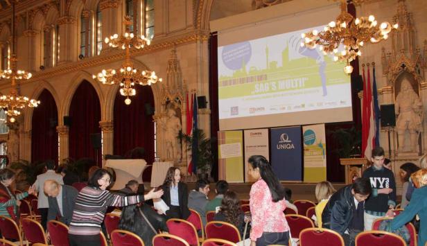 Menschen sitzen in einem Saal mit einer Leinwand, auf der „Sag’s Multi!“ steht.