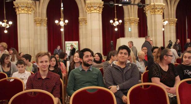 Zuschauer sitzen in einem Saal mit rotem Vorhang und goldenen Säulen.