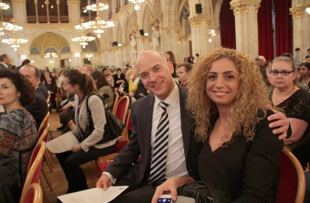 Ein Mann im Anzug und eine Frau sitzen lächelnd in einem festlichen Saal voller Menschen.