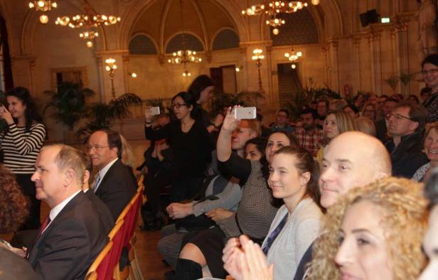 Ein Publikum sitzt in einem opulenten Saal und einige Personen machen Fotos mit ihren Smartphones.