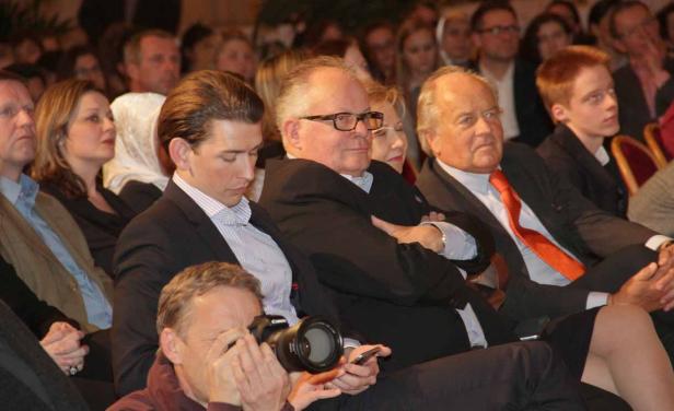 Mehrere Personen sitzen in einer Menschenmenge, darunter ein Fotograf im Vordergrund.