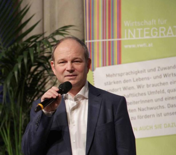 Ein Mann in einem Anzug spricht in ein Mikrofon vor einem Banner der „Wirtschaft für Integration“.