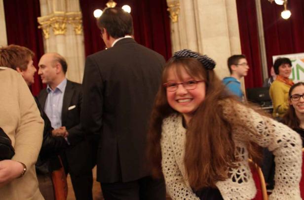 Ein lächelndes Mädchen mit Brille und Haarband steht in einem Raum mit anderen Personen.