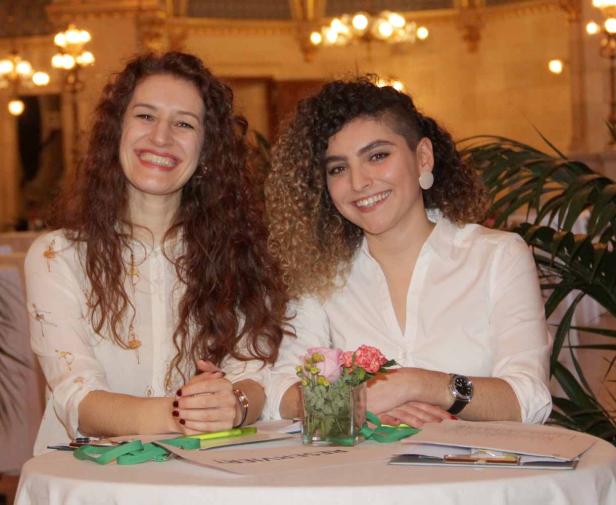 Zwei lächelnde Frauen sitzen an einem Tisch mit Blumen und Unterlagen.