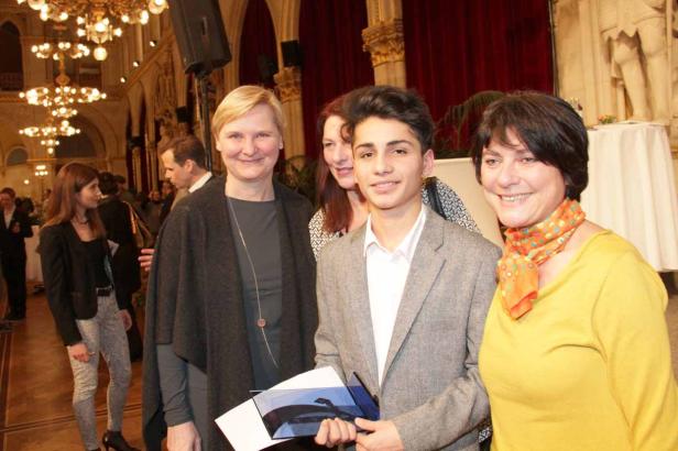 Ein junger Mann hält eine Auszeichnung, umgeben von drei Frauen in einem festlichen Saal.