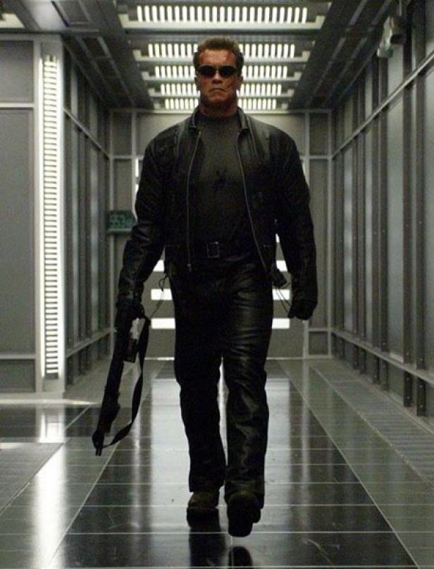 Arnold Schwarzenegger als Terminator geht einen Gang entlang und trägt eine Waffe.