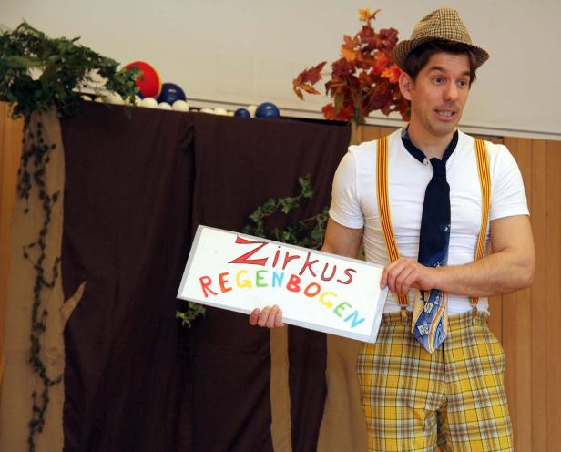 Ein Mann im Clownskostüm hält ein Schild mit der Aufschrift „Zirkus Regenbogen“.