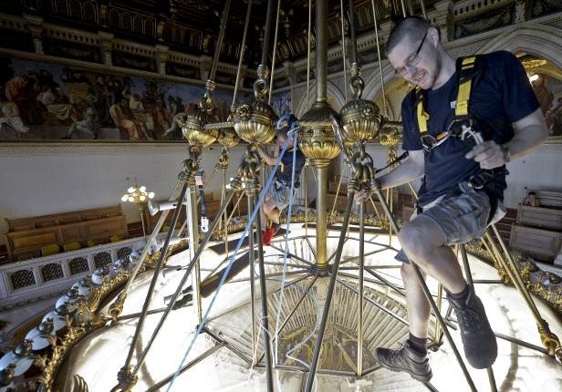 Zwei Männer mit Sicherheitsgurten arbeiten an einem großen, goldenen Kronleuchter in einem verzierten Gebäude.