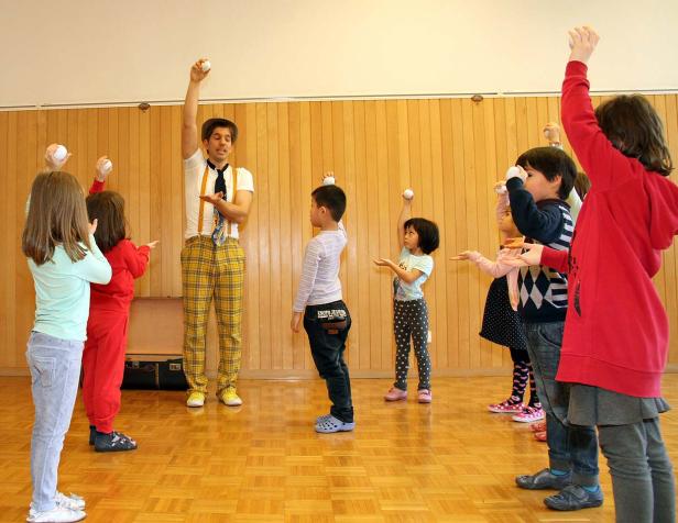 Ein Mann in gelber Hose zeigt einer Gruppe von Kindern, wie man einen Ball hochwirft.
