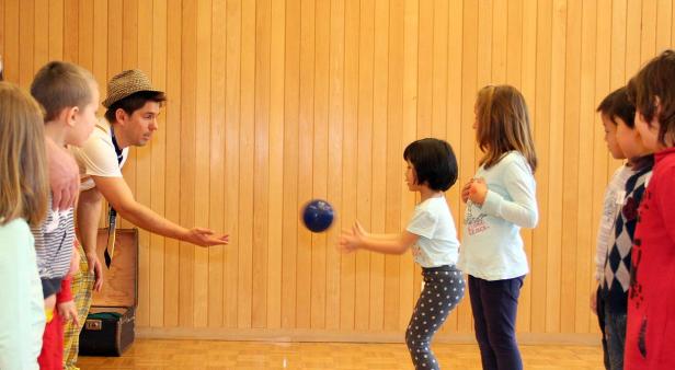 Ein Mann wirft einen blauen Ball zu einem Mädchen vor einer Gruppe von Kindern.