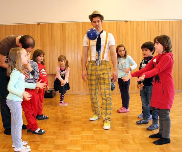 Ein Clown balanciert einen blauen Ball auf seinem Hals vor einer Gruppe von Kindern.