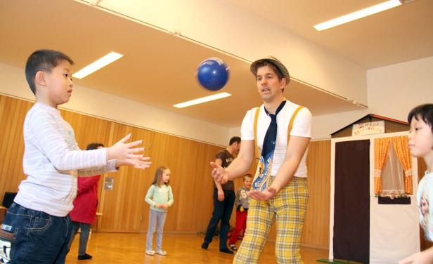 Ein Clown jongliert mit einem blauen Ball vor einer Gruppe von Kindern.