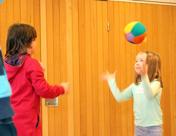 Zwei Kinder spielen mit einem bunten Ball vor einer Holzwand.