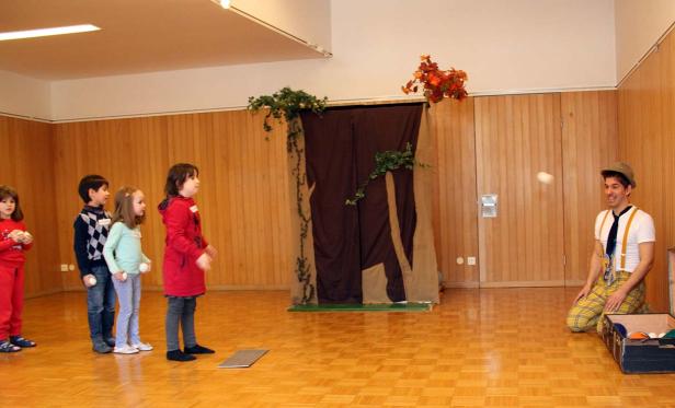 Ein Clown jongliert mit Bällen vor einer Gruppe von Kindern in einem Raum mit Holzwänden.