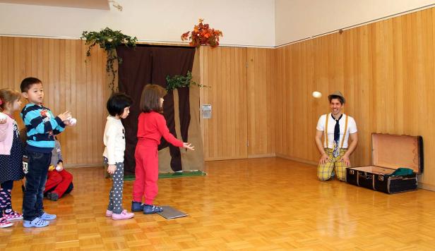 Ein Clown jongliert vor einer Gruppe von Kindern in einem Raum mit Holzboden.