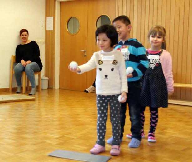 Eine Gruppe von Kindern steht bereit, um mit weißen Bällen zu werfen, während eine Frau zusieht.