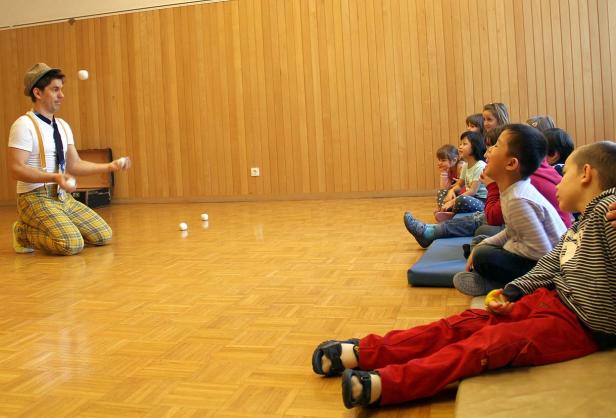 Ein Clown jongliert vor einer Gruppe von Kindern in einem Raum.